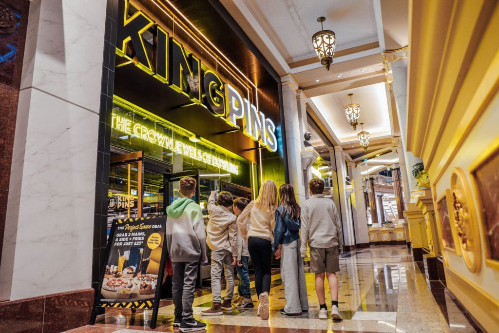 A group of children in a grand building looking up at a florescent sign which says 'King Pins'.